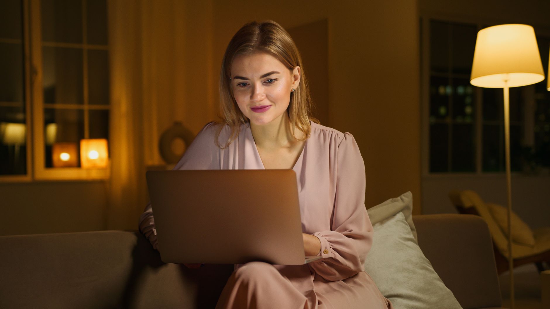Une femme souriante travaille sur un ordinateur portable, installée sur un canapé dans un salon chaleureux, en soirée.